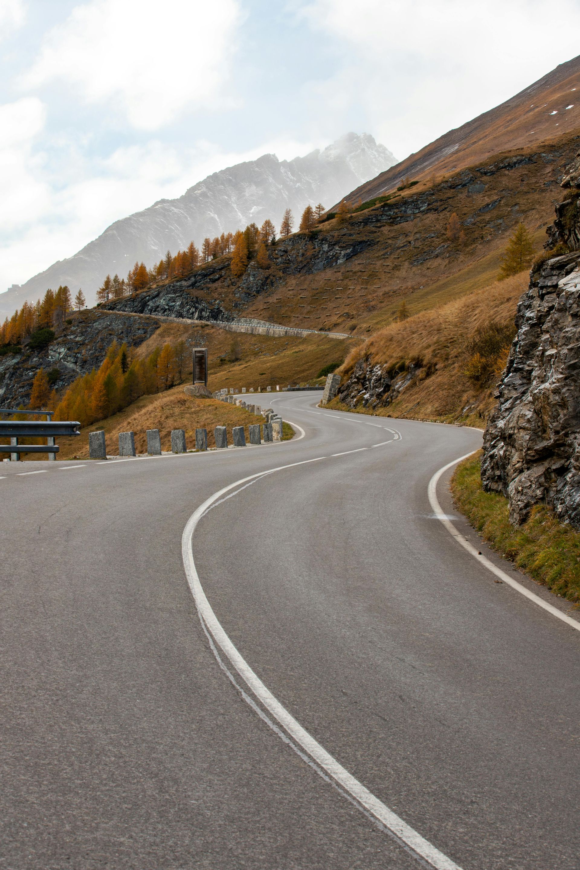Winding mountain road with a snowy peak in the distance, autumn colors visible on the hillside.