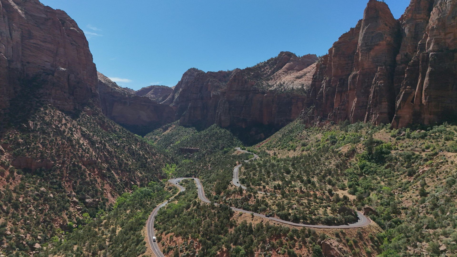 Winding road through a canyon with red rock walls and green trees under a blue sky.
