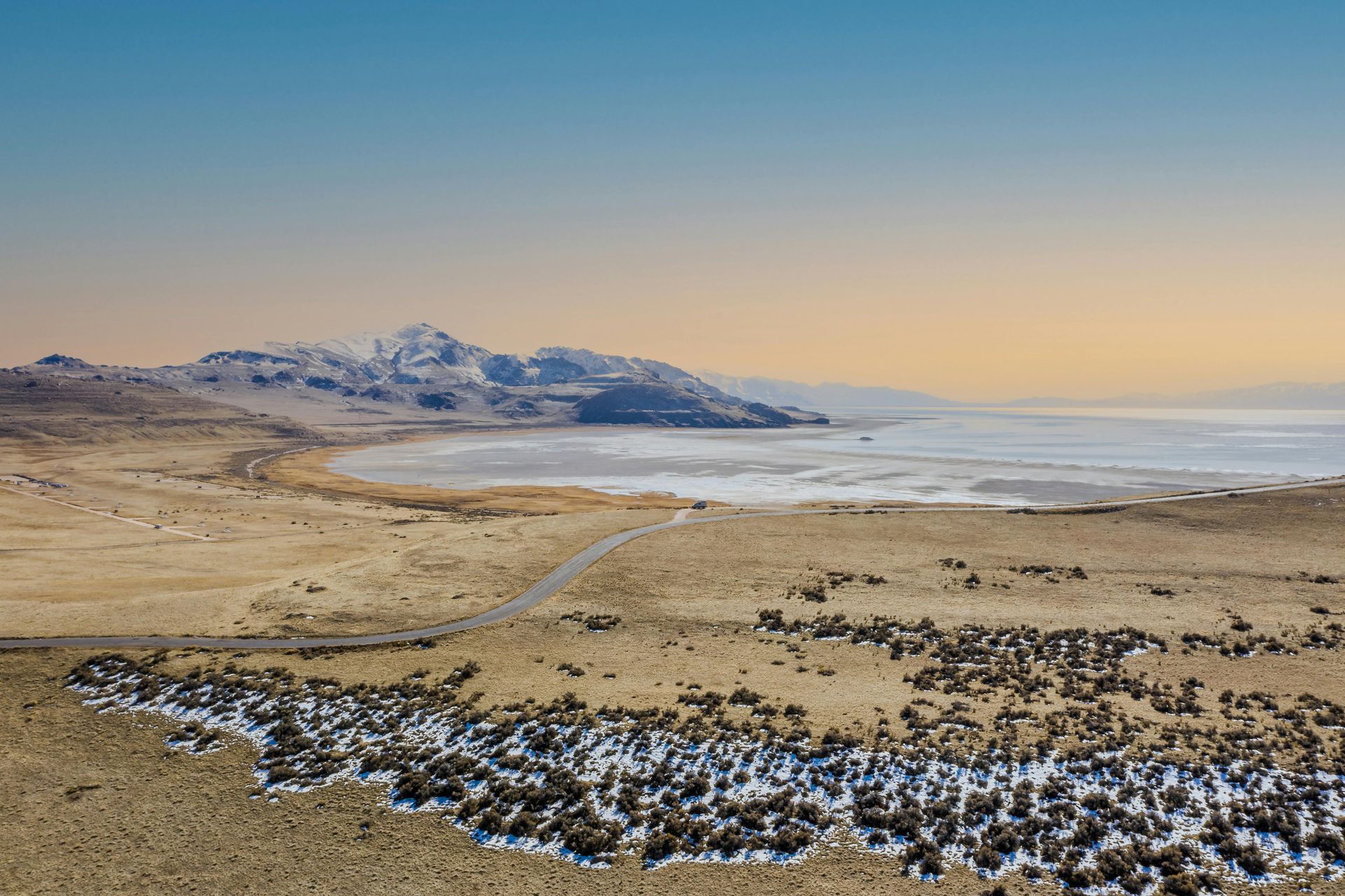 Desert landscape with road leading to a lake, mountains in the distance, and blue sky.