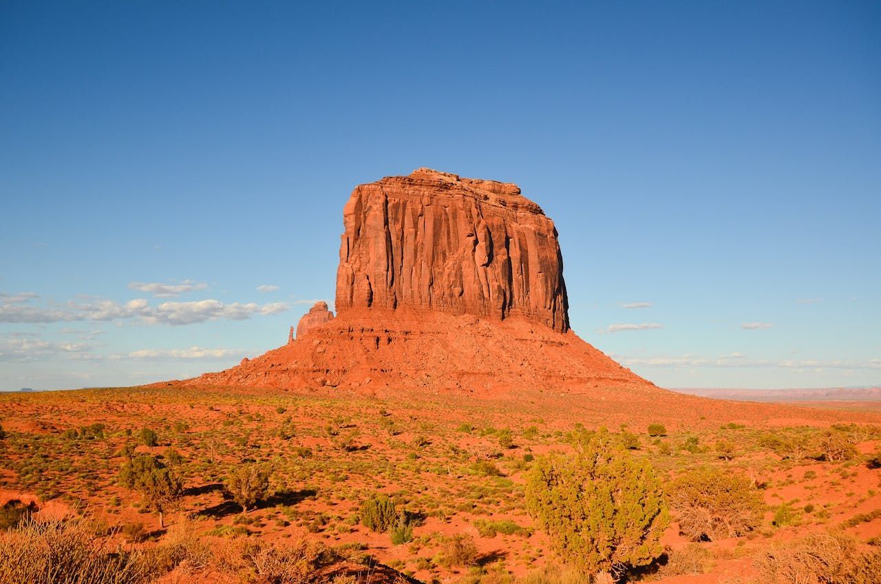 Red sandstone butte against a blue sky, in a desert landscape with sparse vegetation.