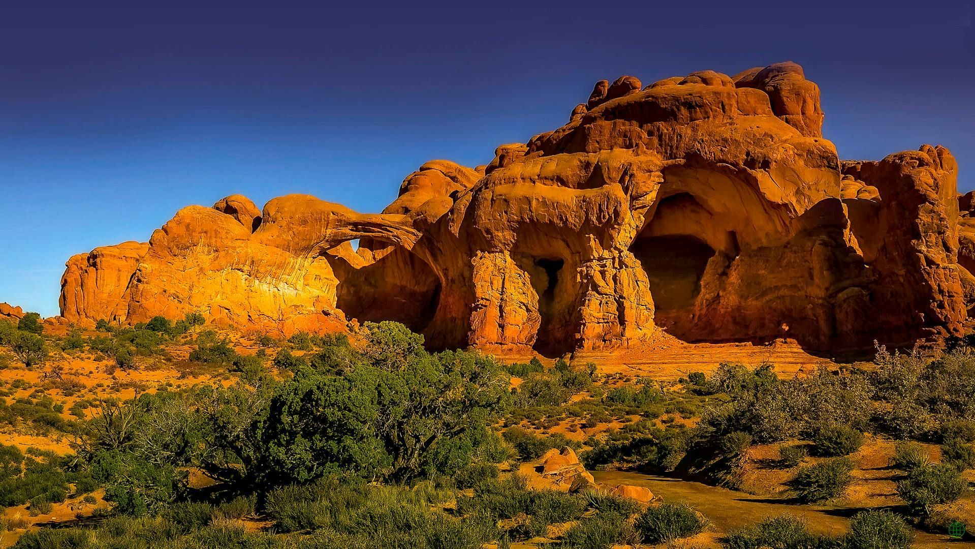 Orange sandstone rock formations under a clear blue sky, with sparse green shrubbery in the foreground.