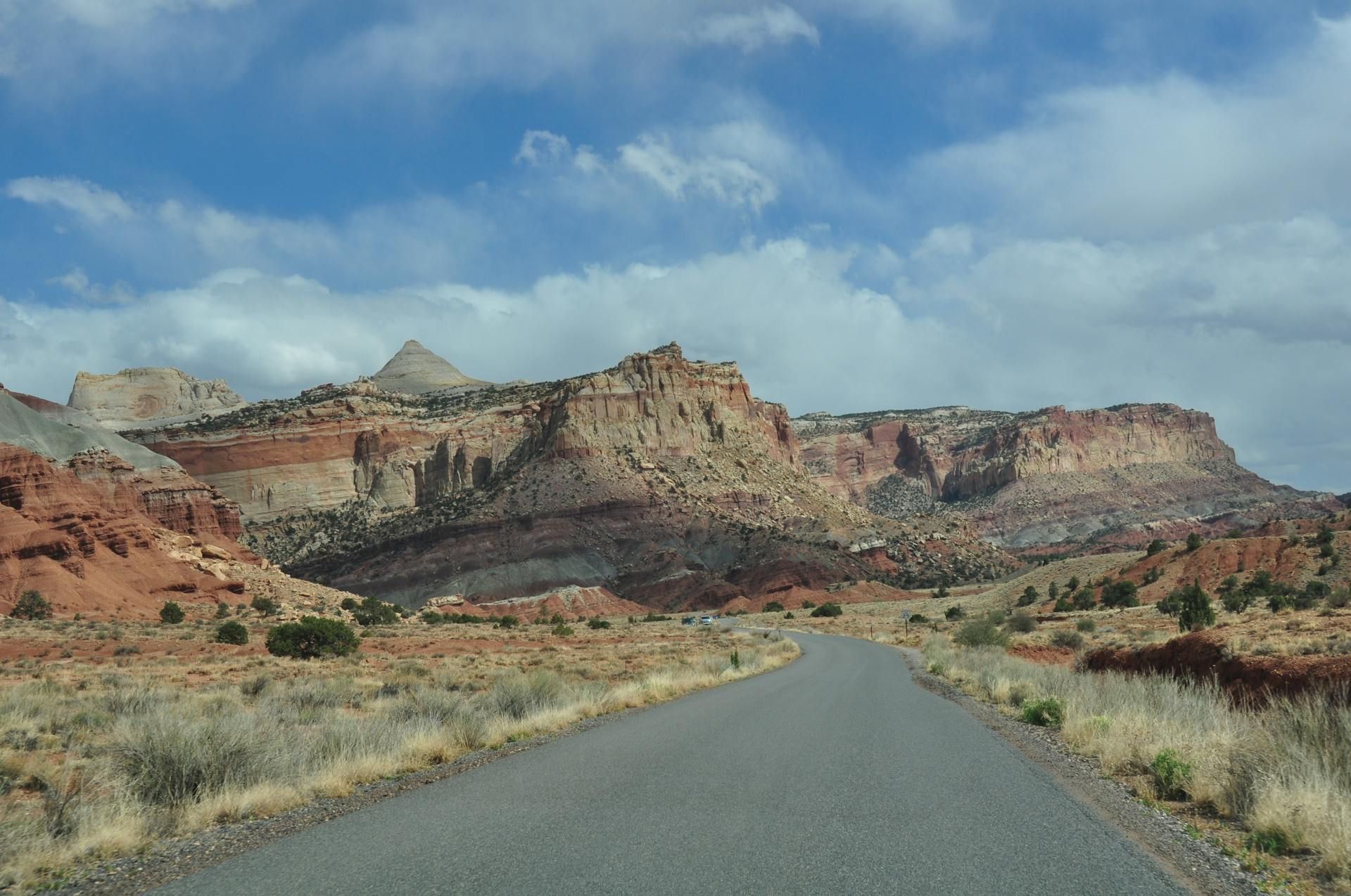 Paved road through a desert landscape; red-rock cliffs under a partly cloudy blue sky.