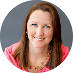 Woman with brown hair and a pink top smiling, wearing a necklace.