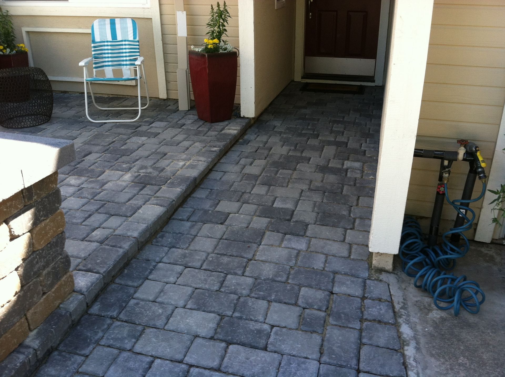 Brick patio leading to a house entrance with a ramp. Blue hose, white chair, and flower pots are visible.