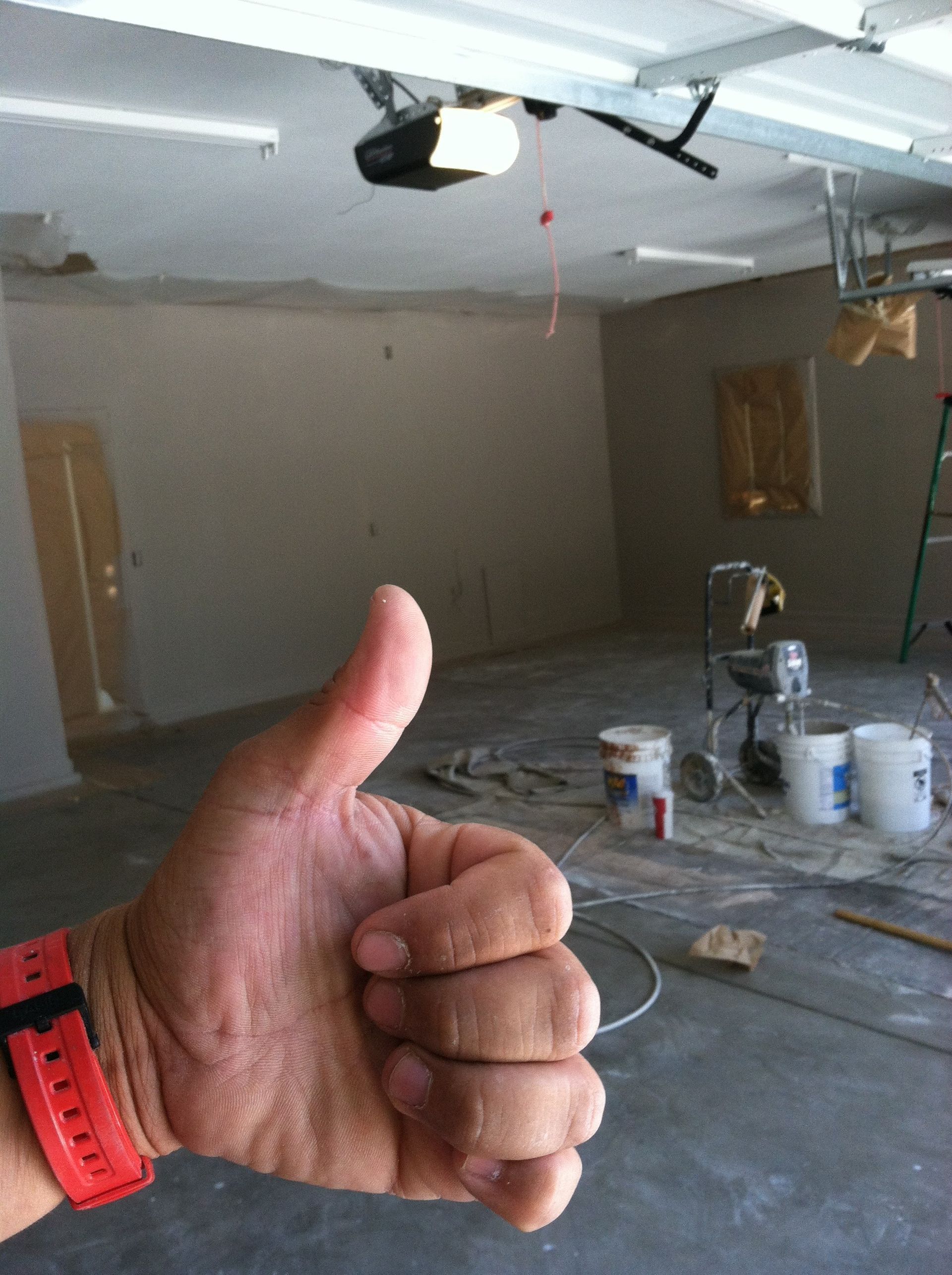 Hand with thumb up in front of an unfinished room; paint buckets and equipment in the background.