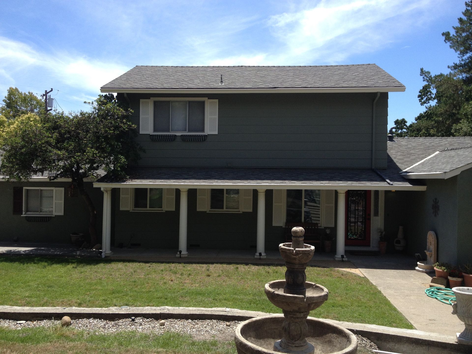 Two-story green house with white trim and a fountain in the front yard under a blue sky.
