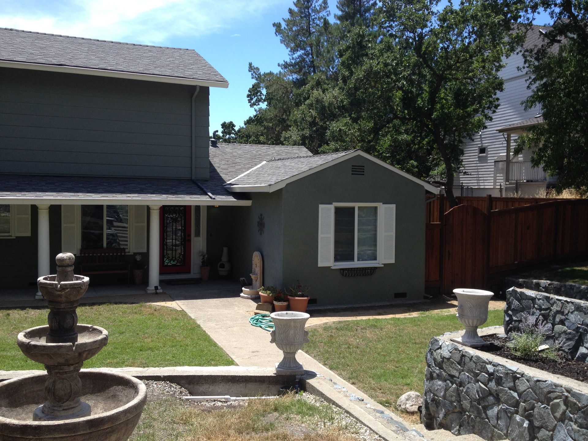 Green house with fountain, small structure, lawn, and wooden fence.