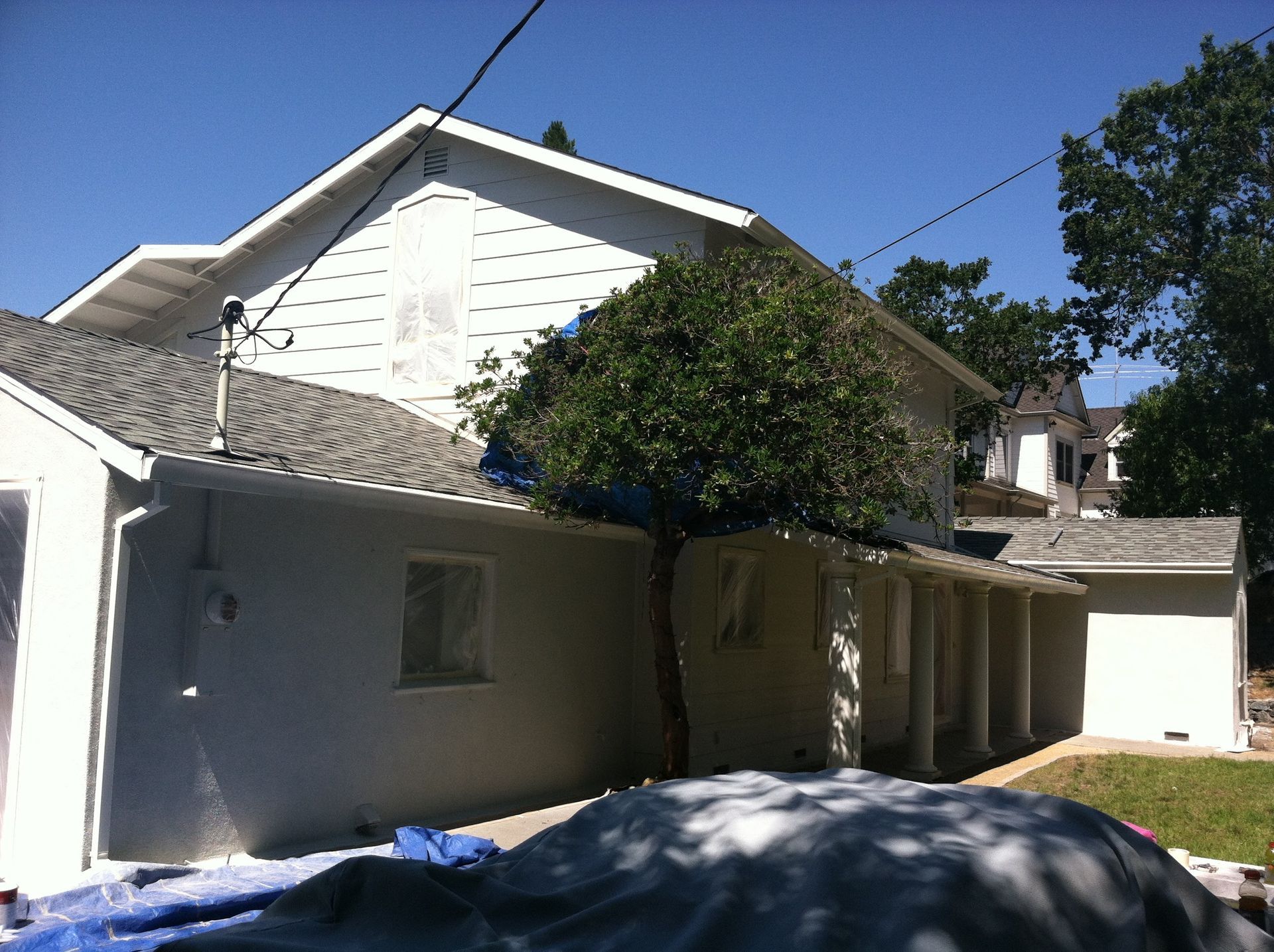 White house with gray roof, a tree growing up the side, and a tarp in the foreground.