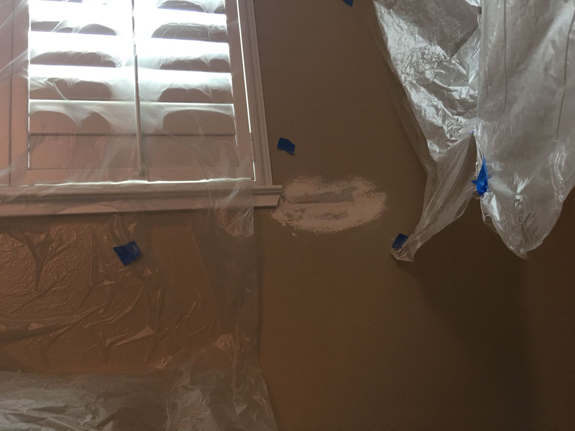 Wall being prepped for painting; plastic sheeting protects window and surrounding areas.