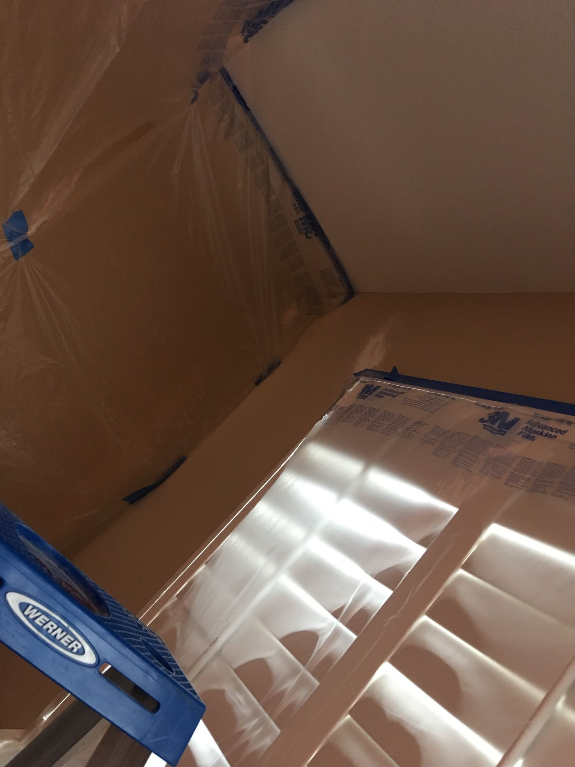 Interior corner of a room, walls and ceiling masked with plastic and blue painter's tape; a window with white shutters is visible.