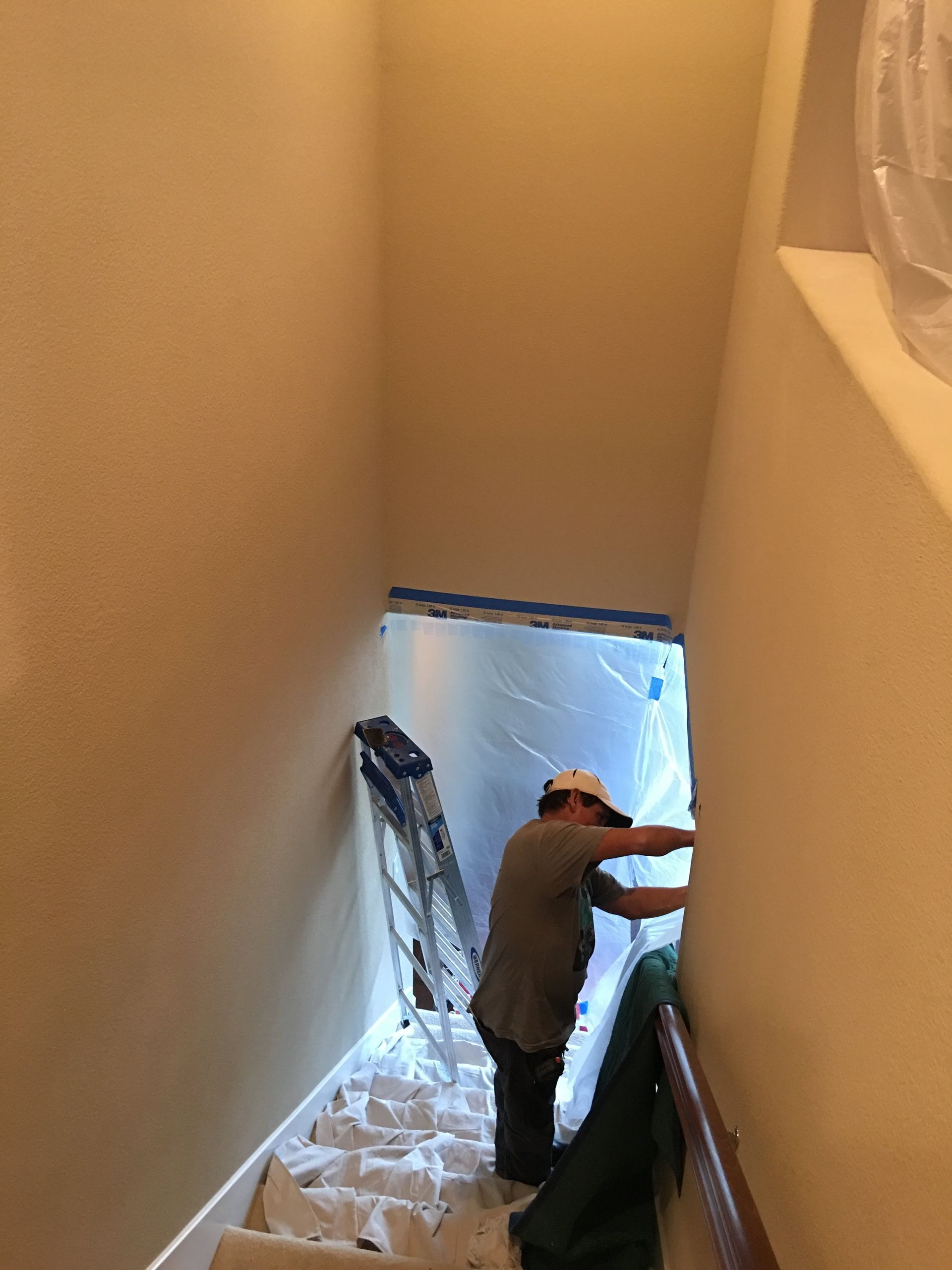 Person on staircase painting a hallway; ladder and drop cloths visible.