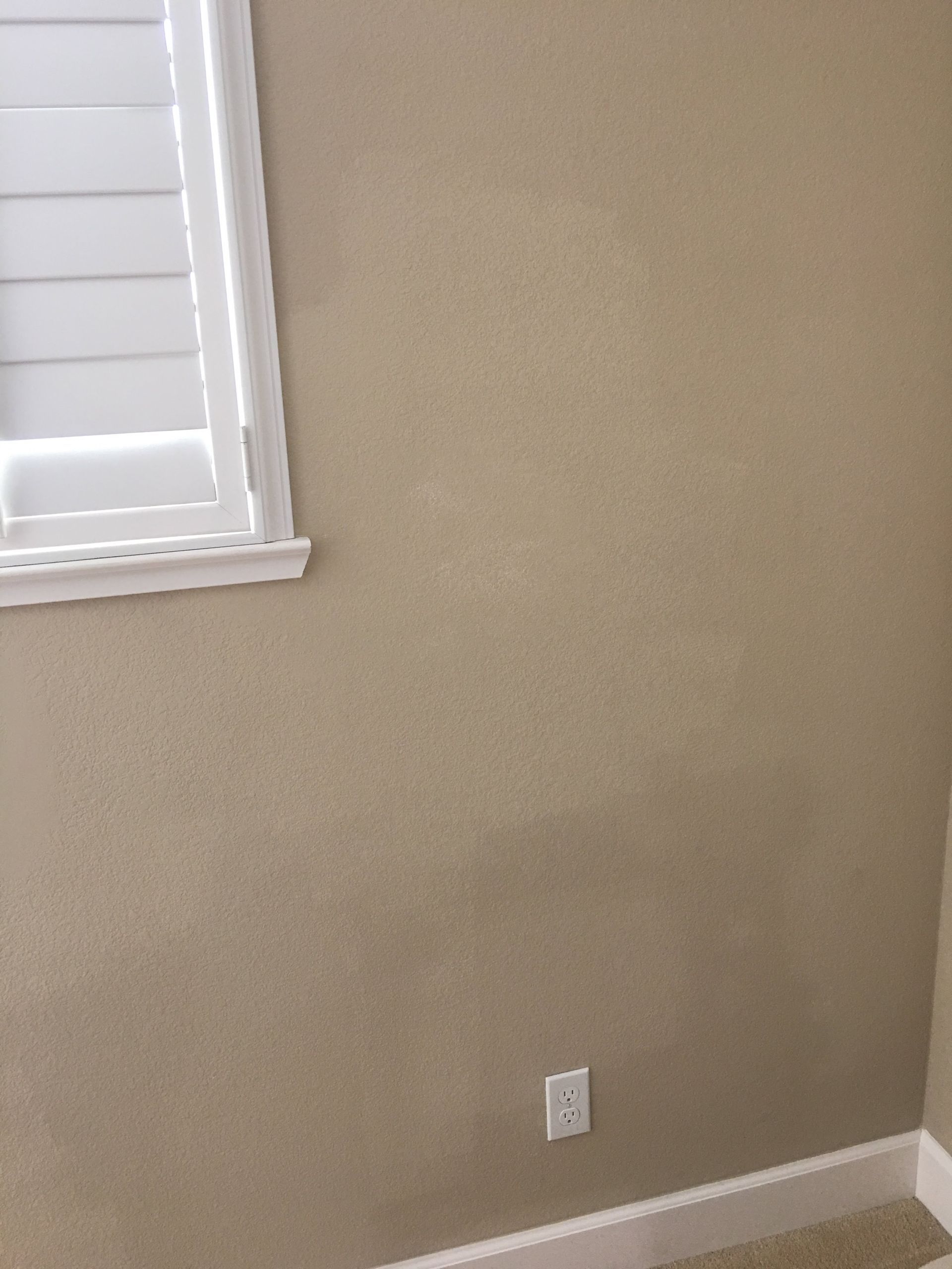 Tan textured wall with a white window and electrical outlet. Beige baseboards.