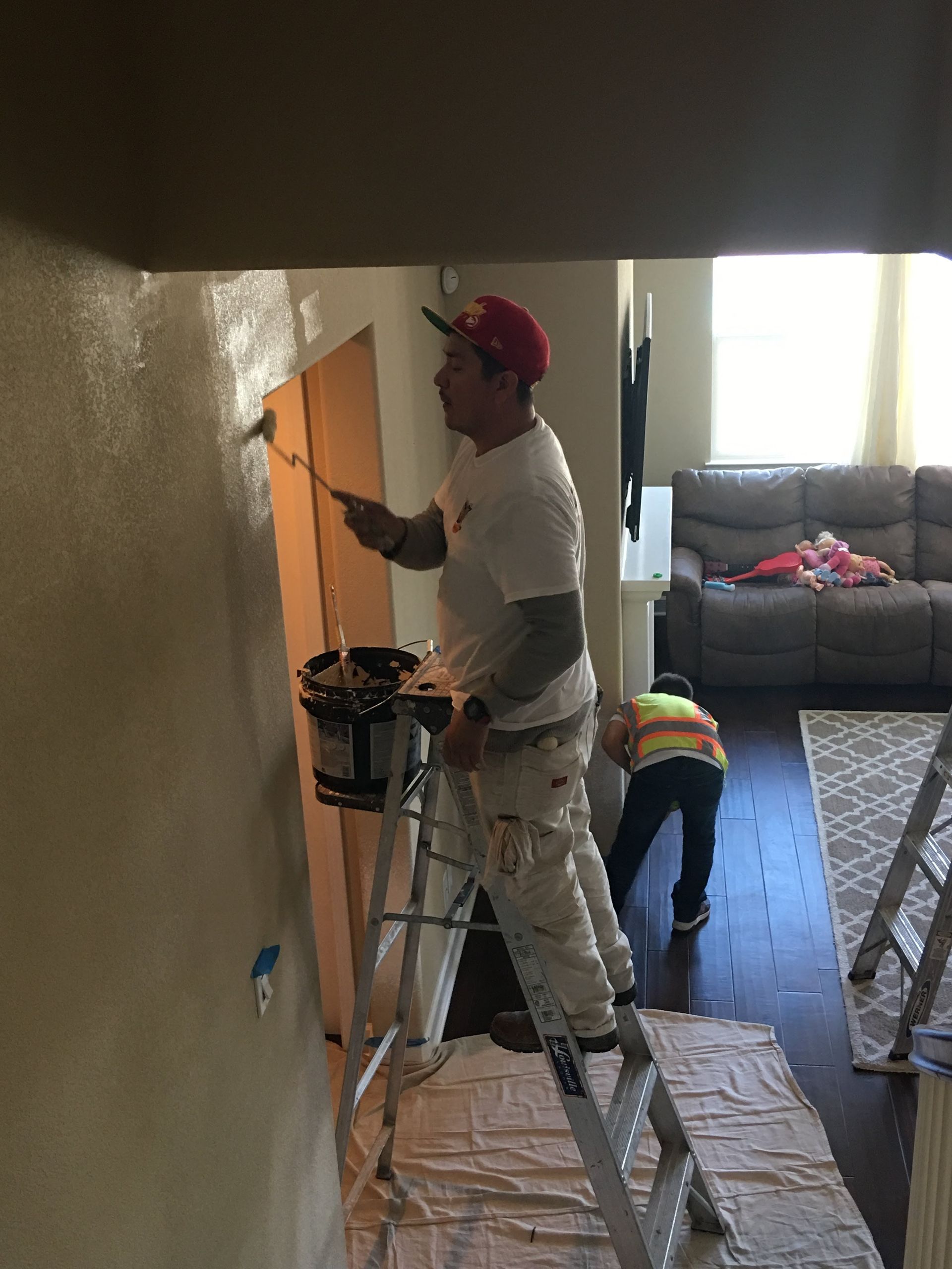 Man in paint-splattered clothes painting a wall. A child watches nearby. Indoors.