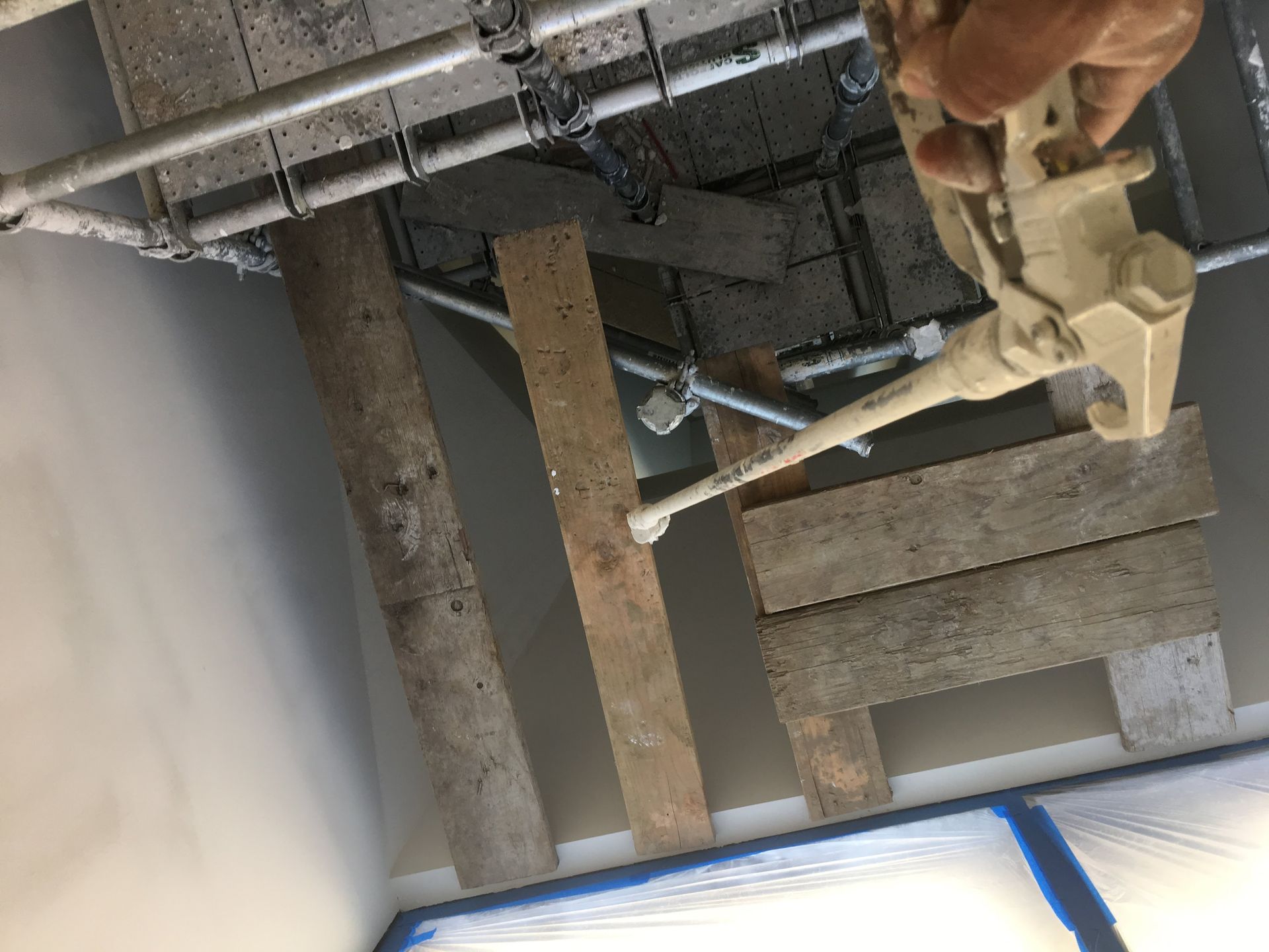 Person applying sealant from a spray gun to ceiling beams. Protective plastic covers nearby walls.