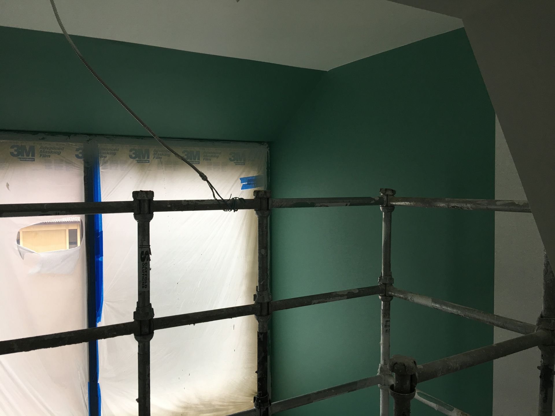 Interior corner with green painted walls, scaffolding, and plastic sheeting.