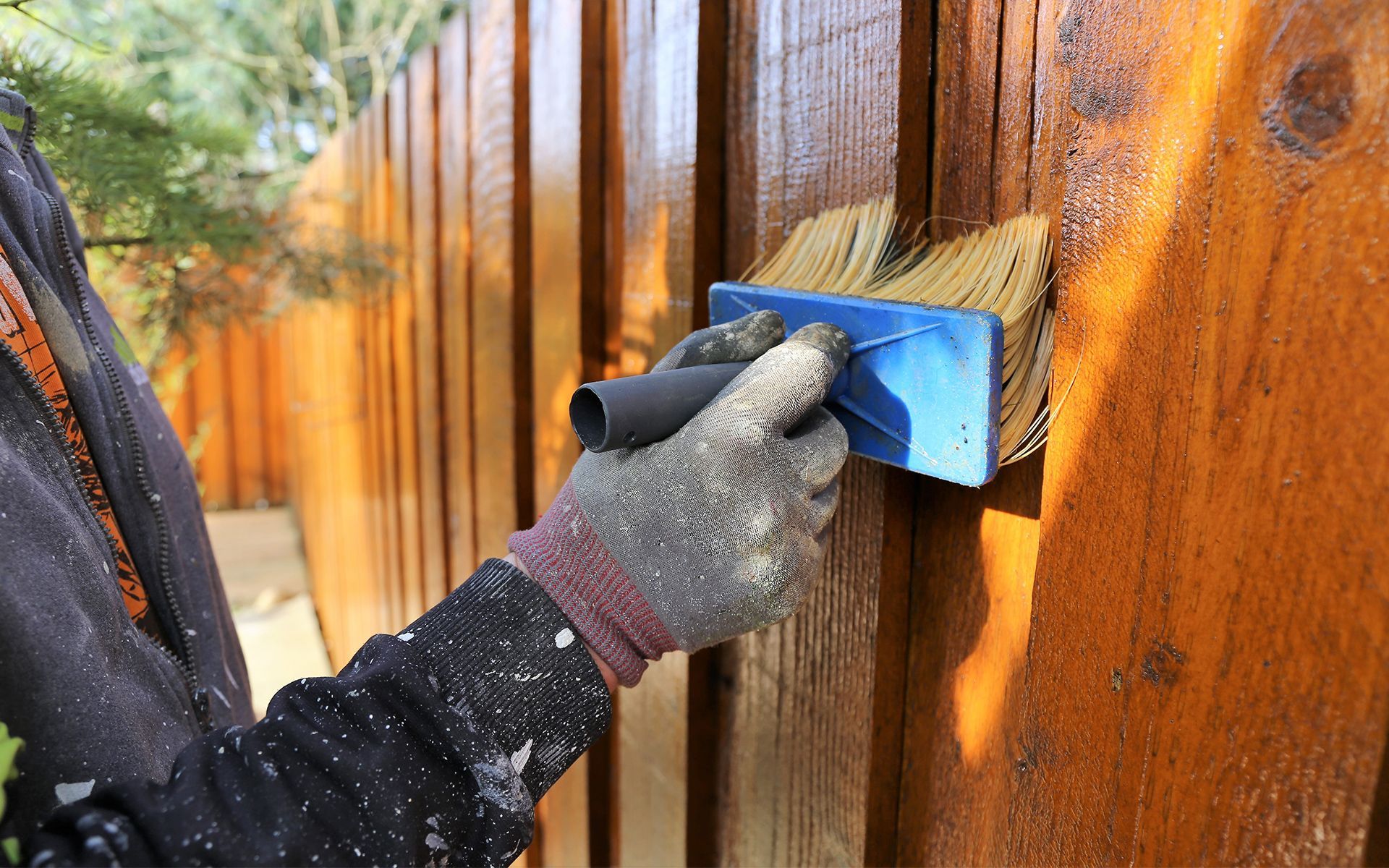 Person staining a wooden fence with a brush, wearing work gloves.