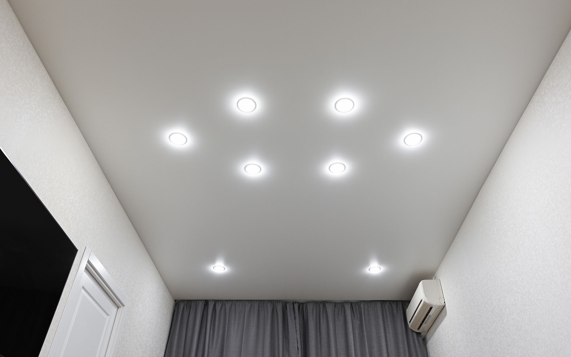 White ceiling with recessed lights, above a room with curtains and an air conditioner.