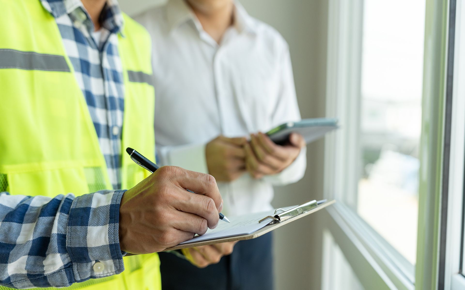 Construction worker in safety vest taking notes while another man holds a tablet indoors.