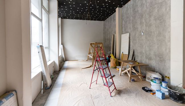 Interior renovation of a room with a red ladder, paint cans, and construction materials; a large window on the left.