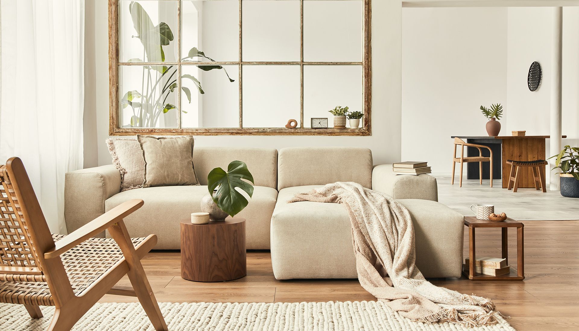 Beige sectional sofa in a bright living room, with a woven chair, plants, and wooden side tables.