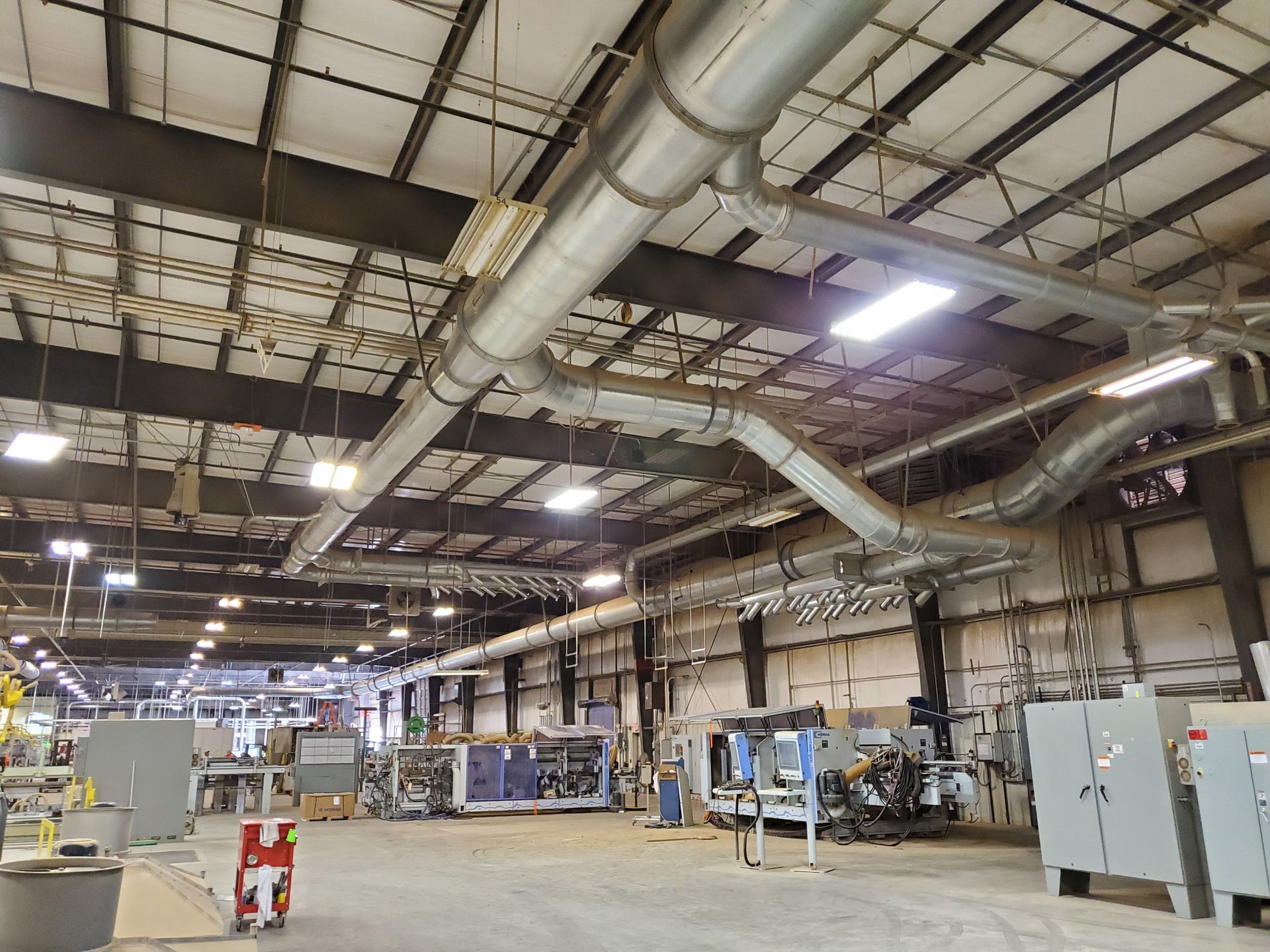 Interior of a high-ceilinged industrial warehouse featuring exposed ductwork, fluorescent lighting, and large machinery.