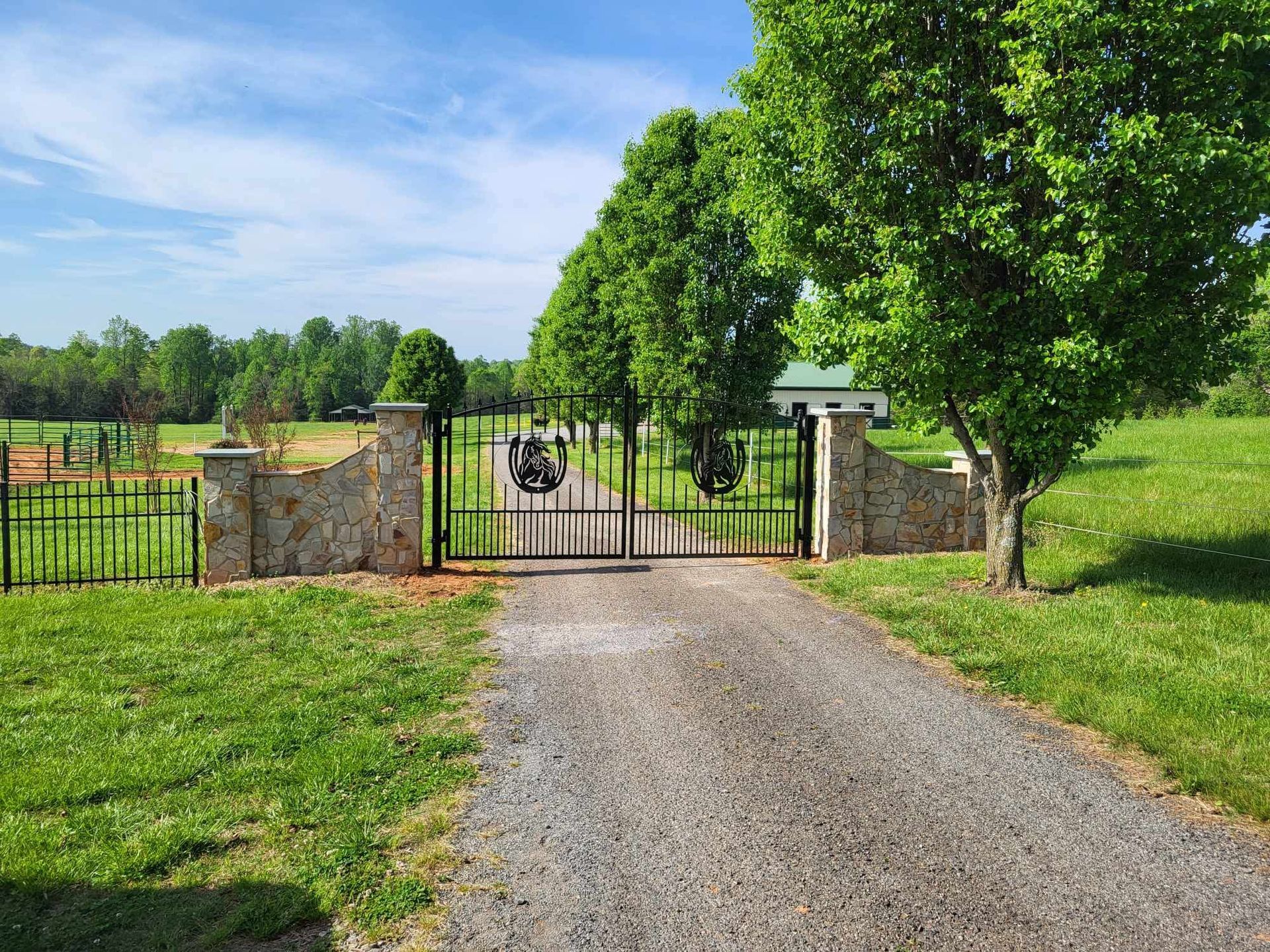 Gravel driveway leading to iron gates flanked by stone walls, trees, and grassy fields under a blue sky.
