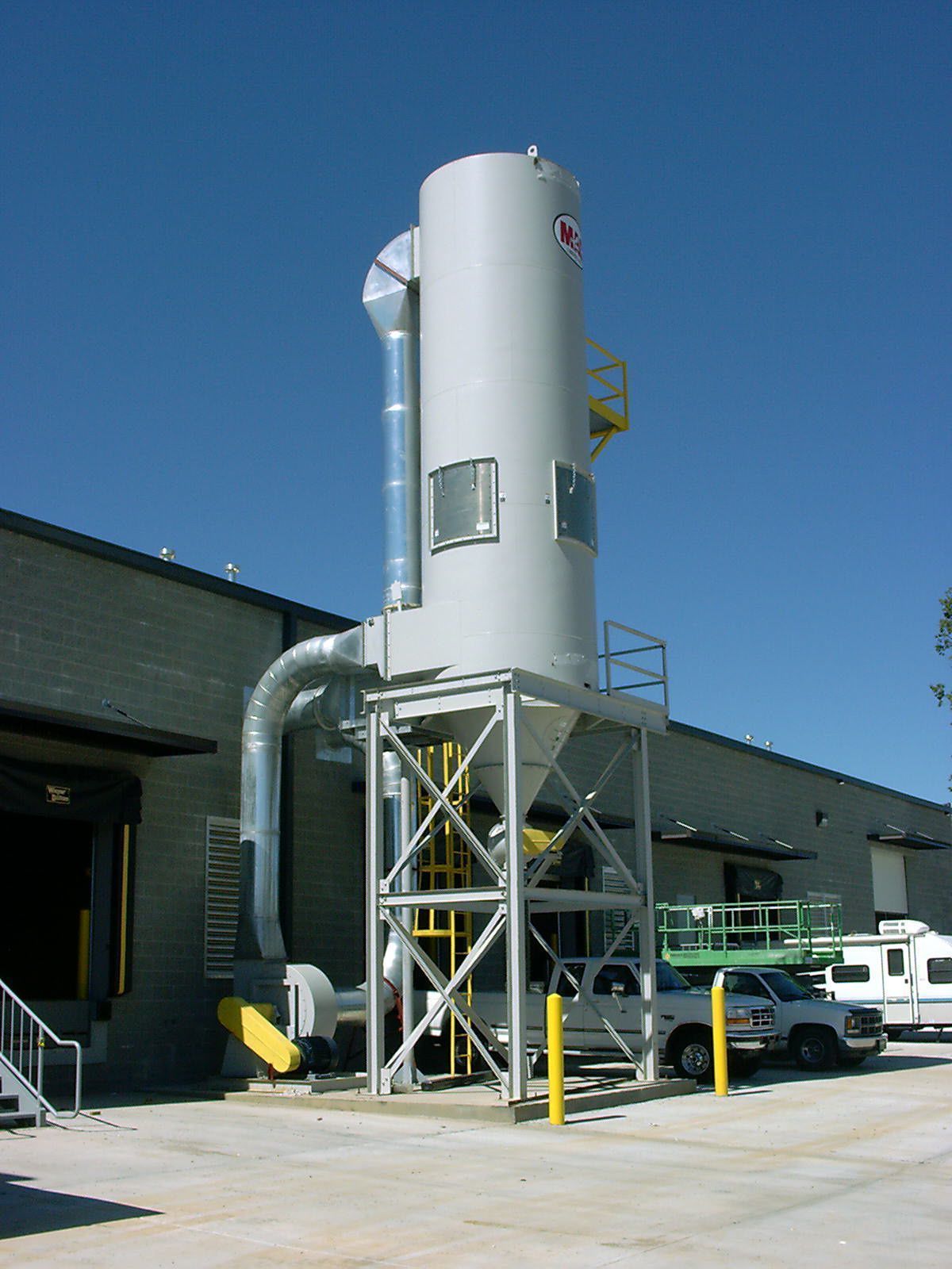 Gray industrial dust collector outside a building, supported by a metal frame. Blue sky.
