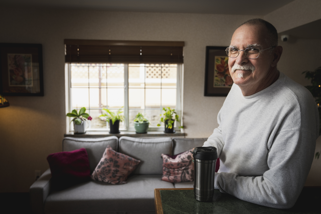 Man with glasses leans near a couch with a coffee mug in a sunlit room.