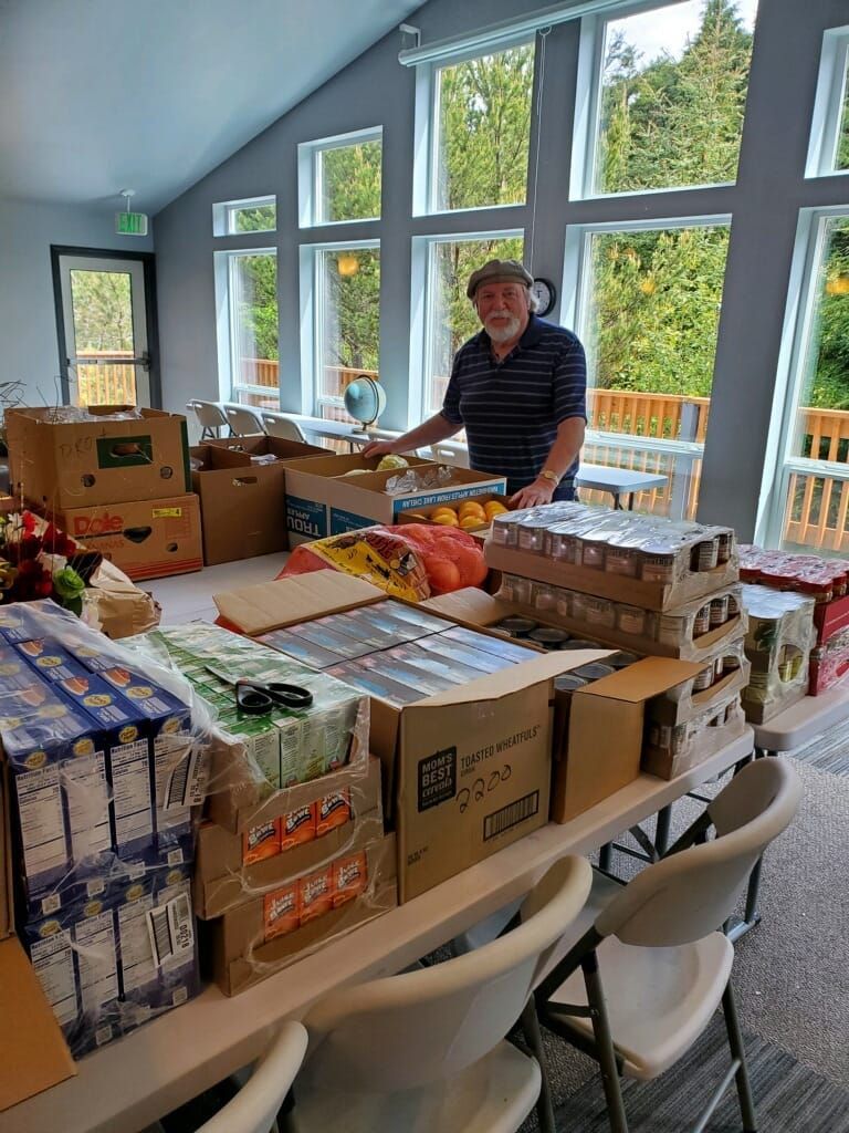 Man preparing food packages at a table with groceries. Inside a bright room with large windows.