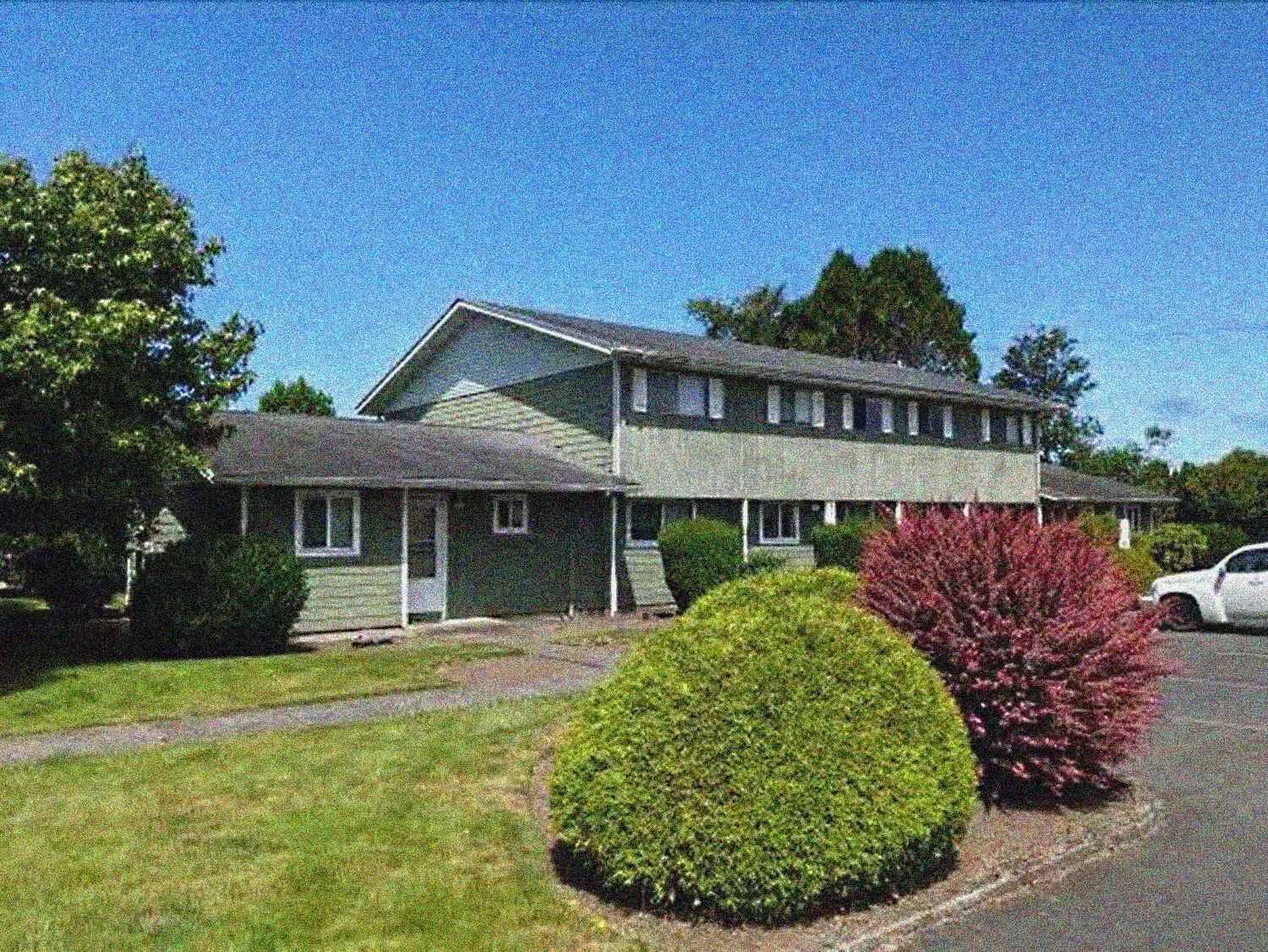 Green two-story building with a flat roof, green lawn, bushes, and a blue sky.