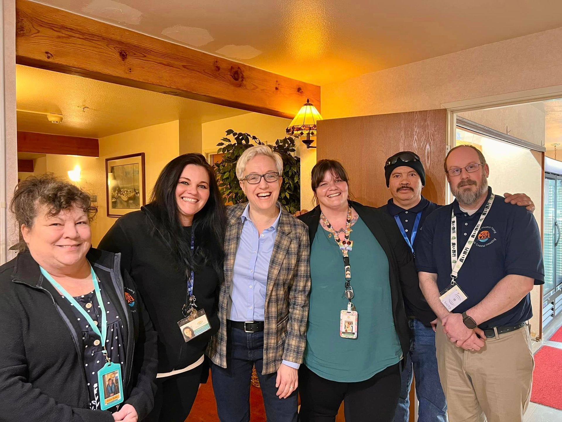 Group of six people smiling indoors; wood beam and open doorway in background.