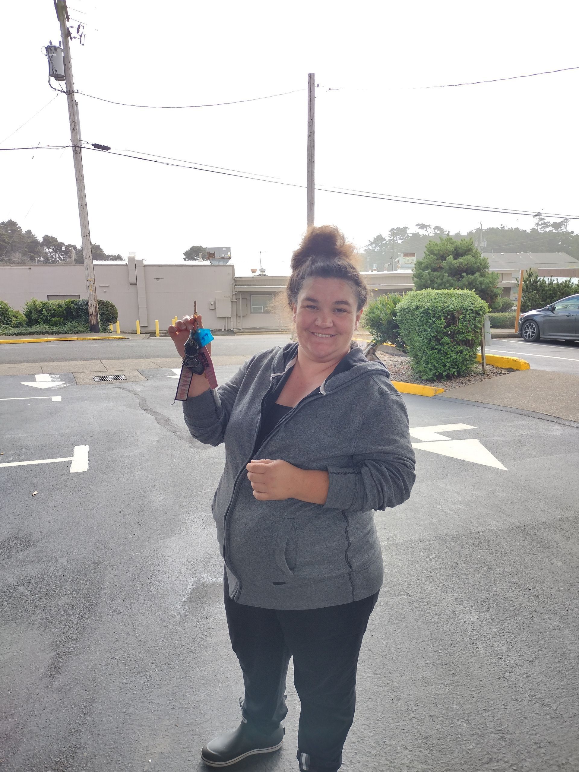 Woman in a gray hoodie holding up keys in a parking lot, smiling.
