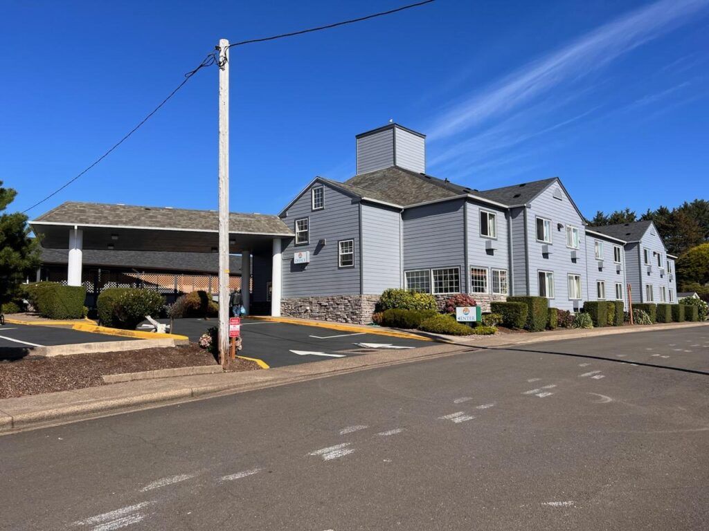 A blue-gray two-story motel with a parking area, under a clear blue sky.