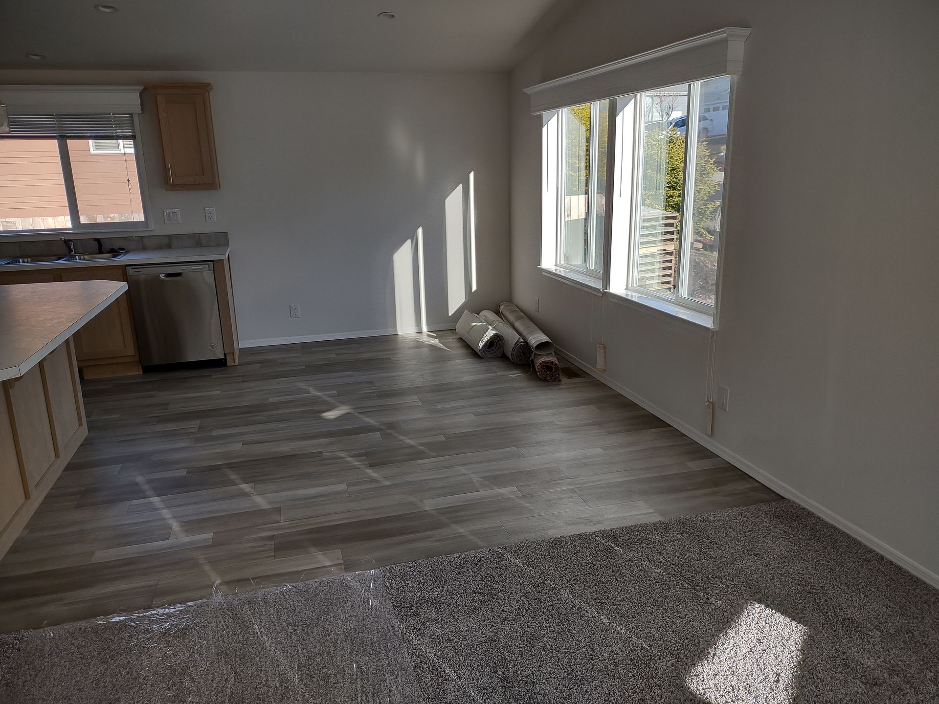 Empty living space with wood-look tile flooring and carpet. Sunlight streams through a window.