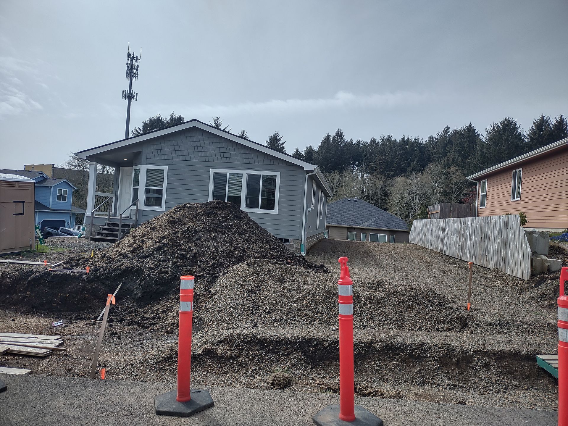 Construction site with gravel piles, orange safety poles, and a small, gray house in the background.