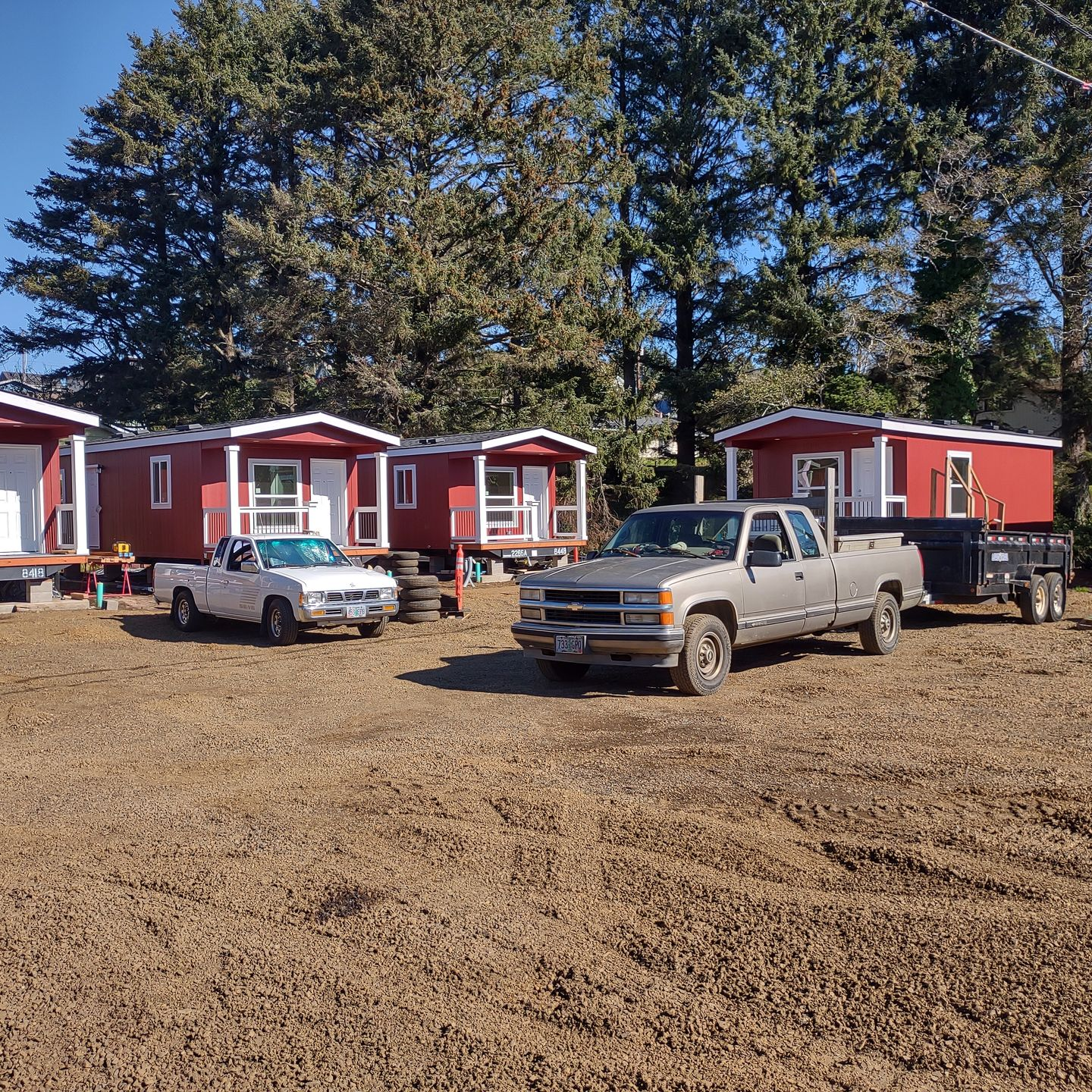 Several small red houses on foundations. Two trucks on gravel, one towing a trailer.