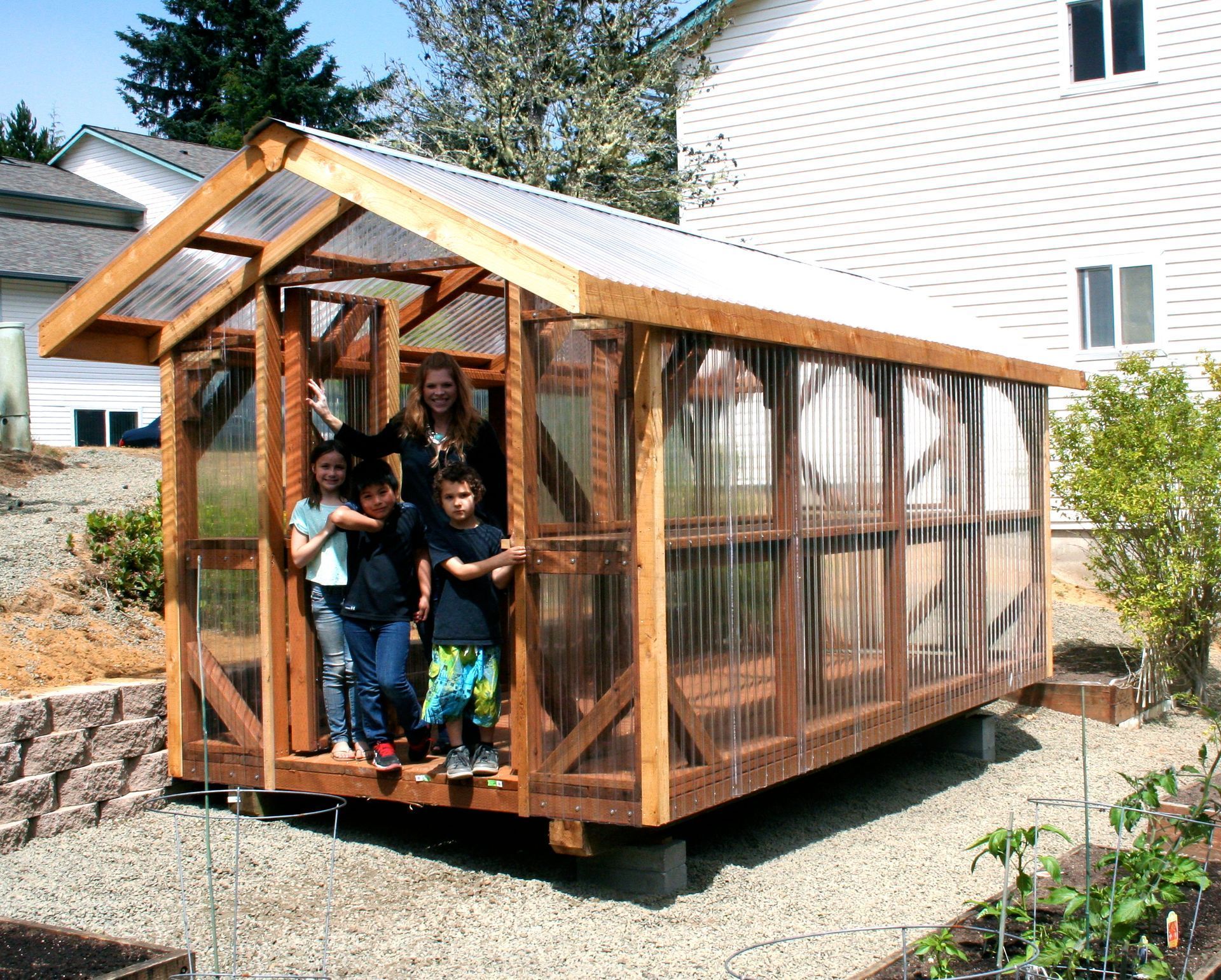Greenhouse with four people standing in the doorway; exterior, wooden frame, clear panels, gravel ground.
