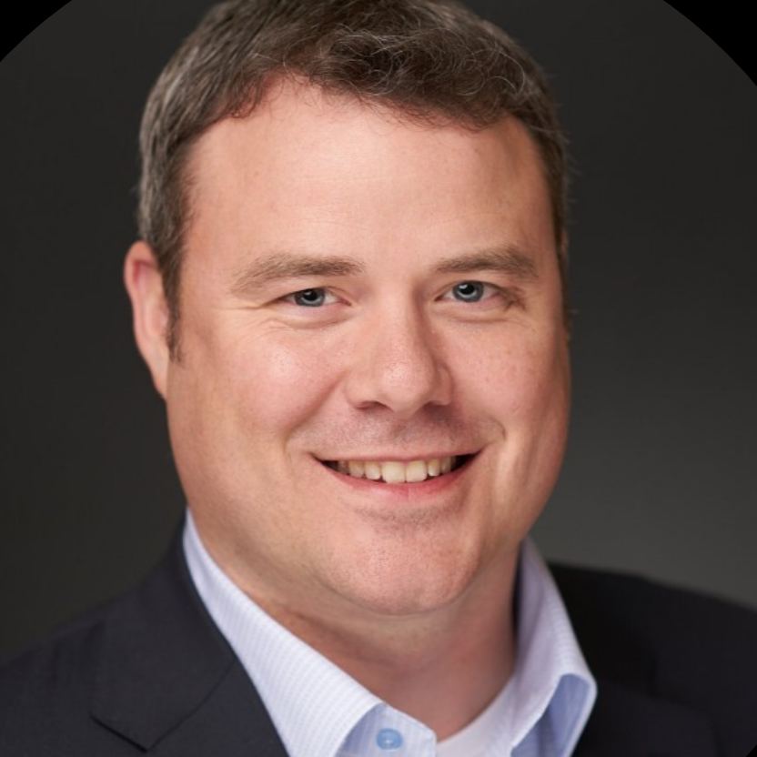 Smiling man in a blue suit and light blue shirt, against a dark background.