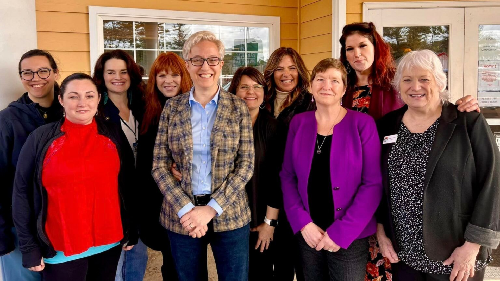 Group of people smiling, standing in front of a building. Many are wearing jackets and looking at the camera.