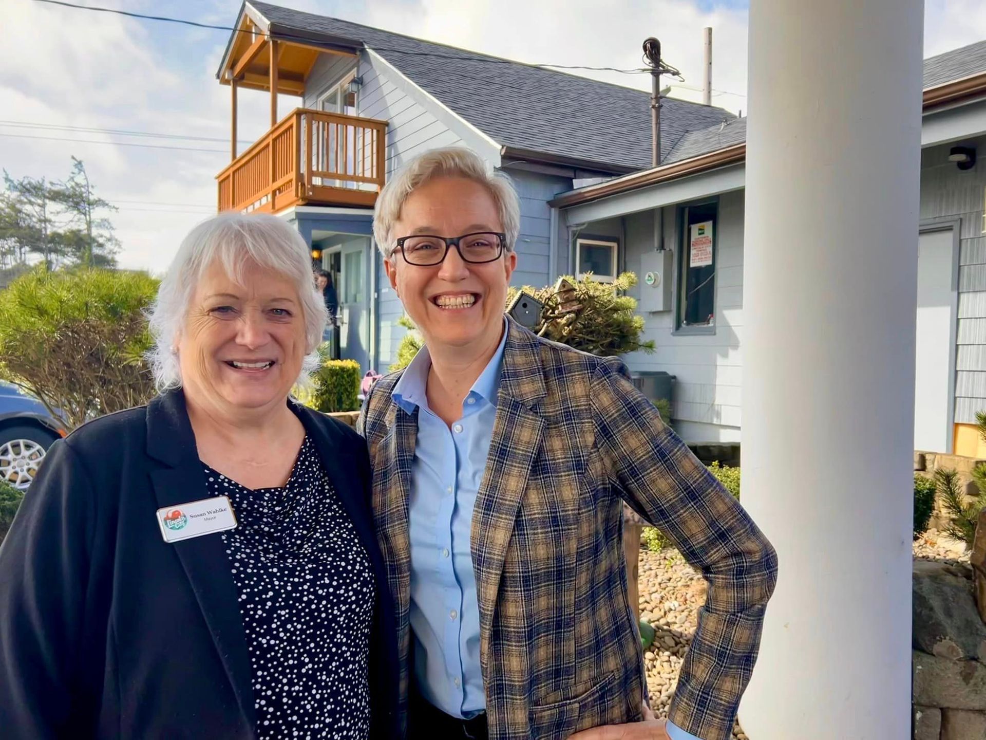 Two smiling people pose outside a building. One wears a patterned blazer, the other a name tag.