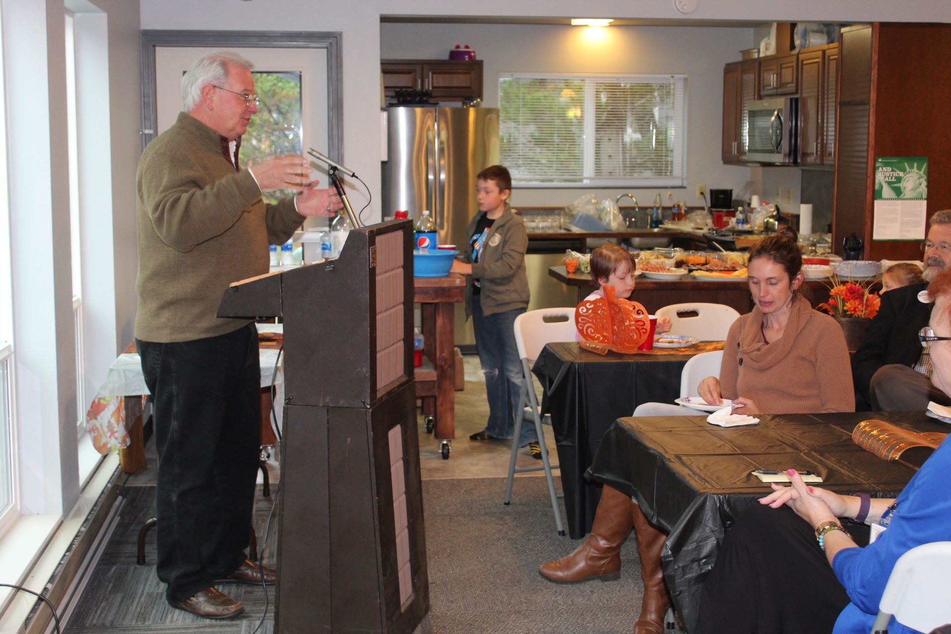 Man speaking at podium in a room, people seated at tables, a boy and kitchen in the background.