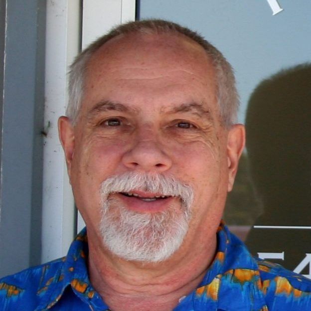 Man with graying hair and goatee smiles at camera, wearing a blue patterned shirt, outdoors.