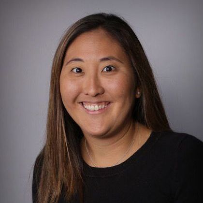 Woman with long brown hair smiling at the camera, wearing a black shirt against a gray background.