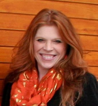 Woman with red hair and scarf smiles in front of a wooden wall.