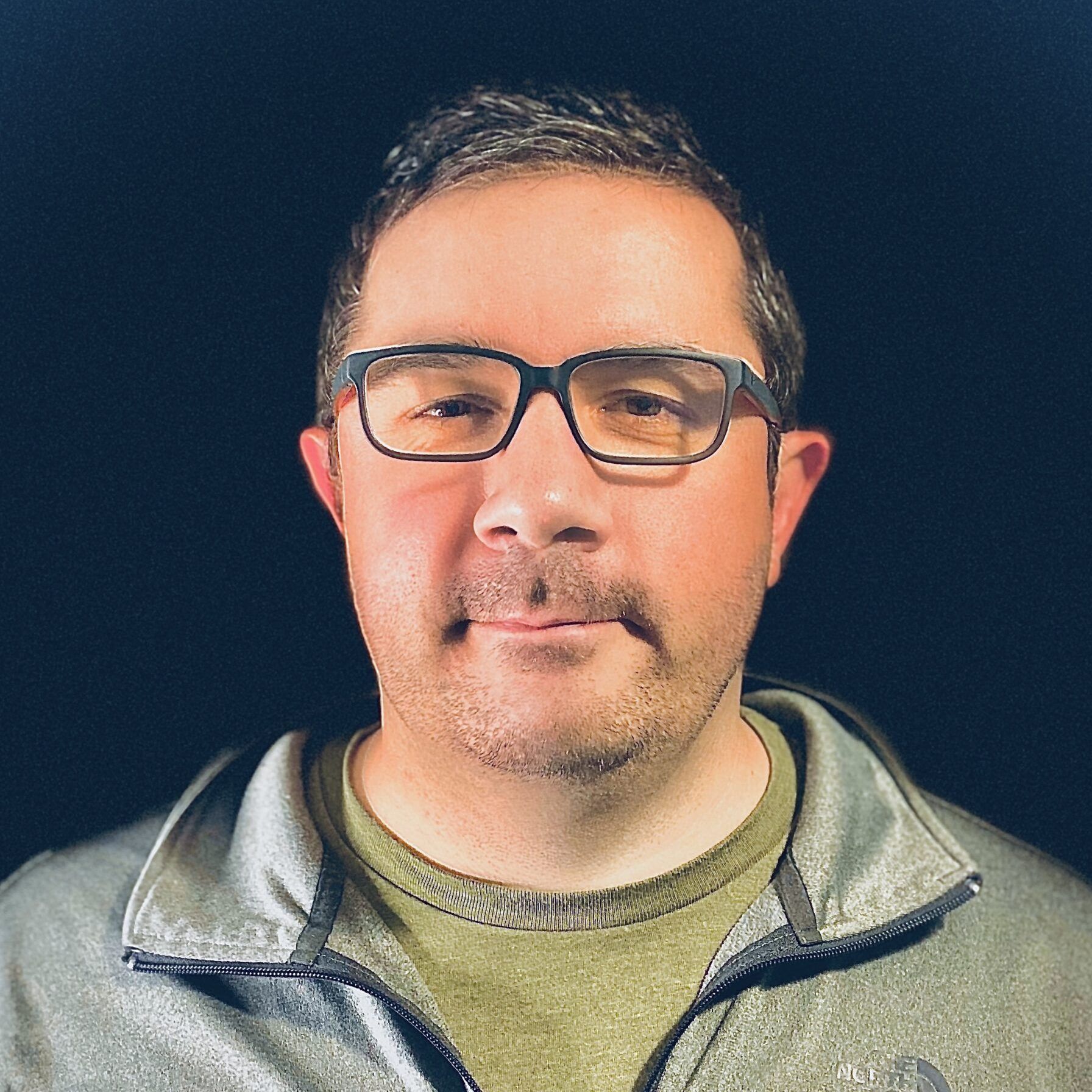 Man wearing glasses, olive green shirt, and gray jacket, looking at the camera. Black background.