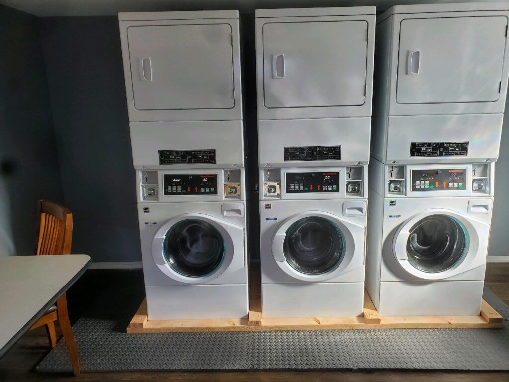 Three stacked white washing machines and dryers in a laundry room; table and chair to the left.