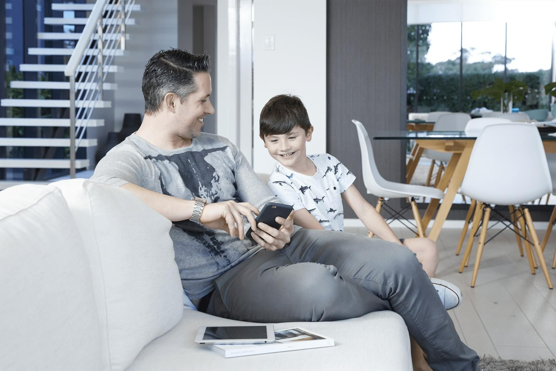 A Man And A Boy Are Sitting On A Couch Looking At A Tablet — Jervis Bay Airconditioning In Huskisson, NSW