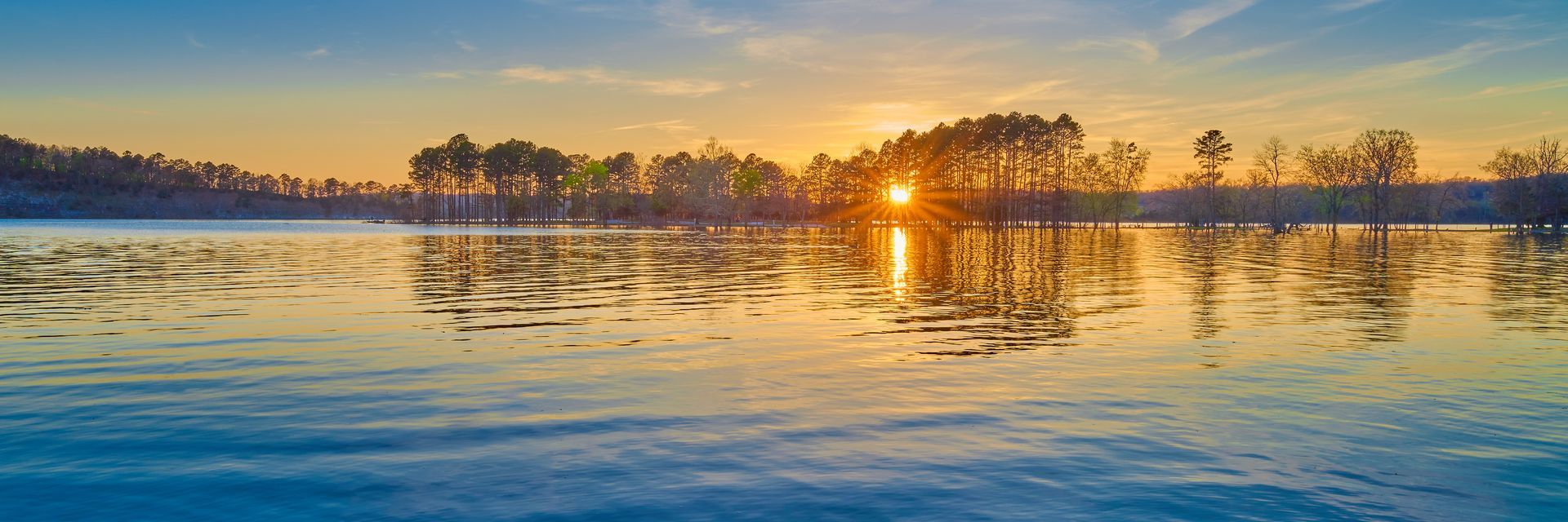 the sun is setting over a lake with trees in the background .