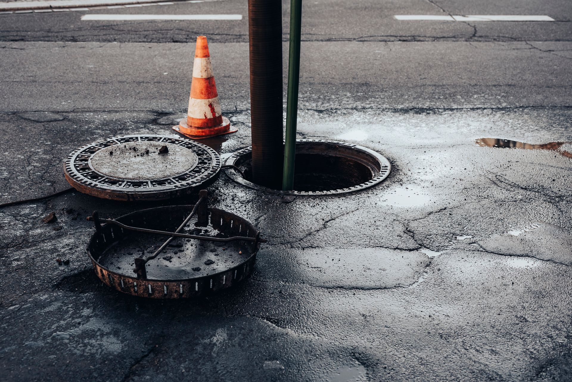 Open sewer access with metal cover and traffic cone on a wet street.