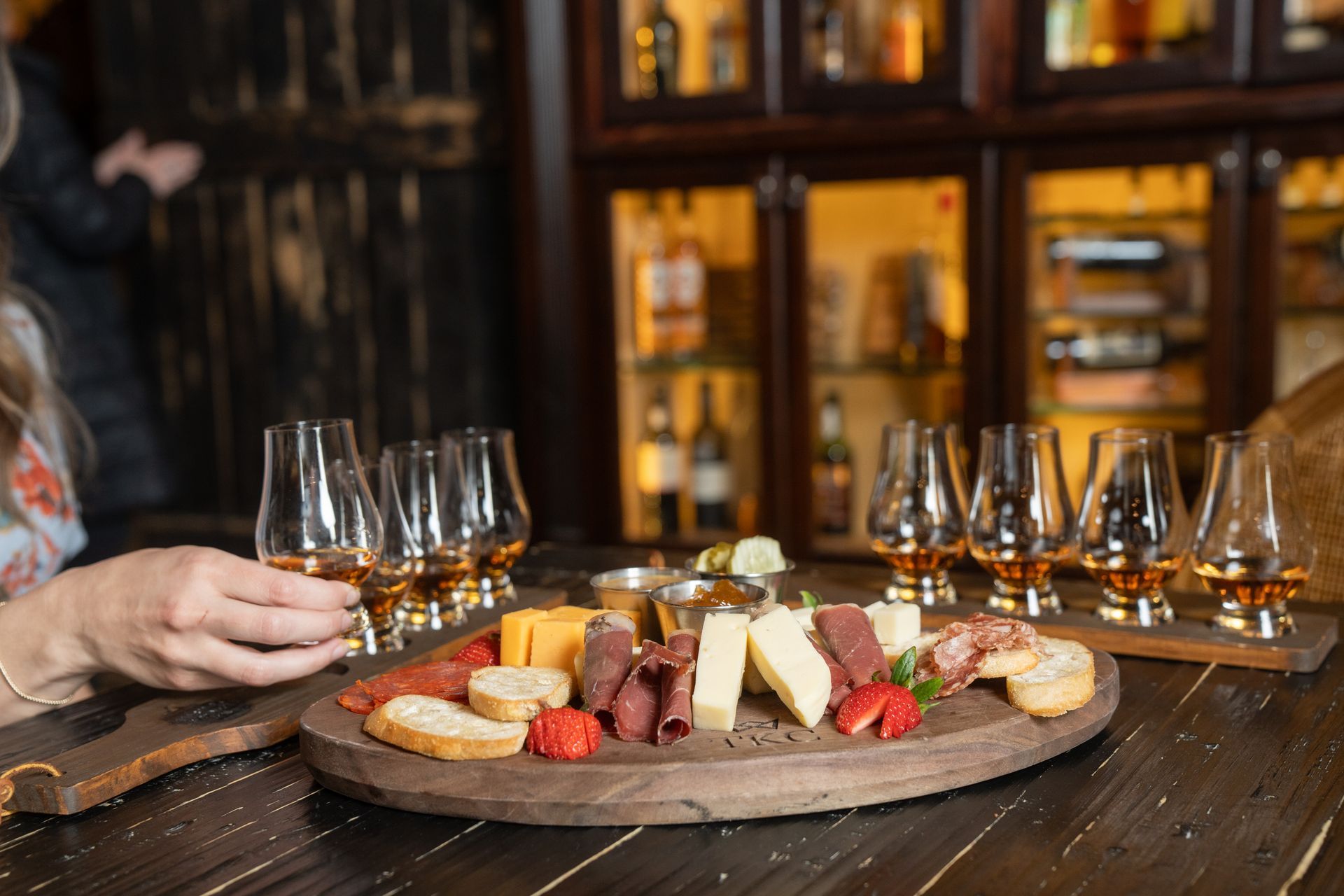 Charcuterie board with whiskey glasses on a dark bar; a person holding a glass.